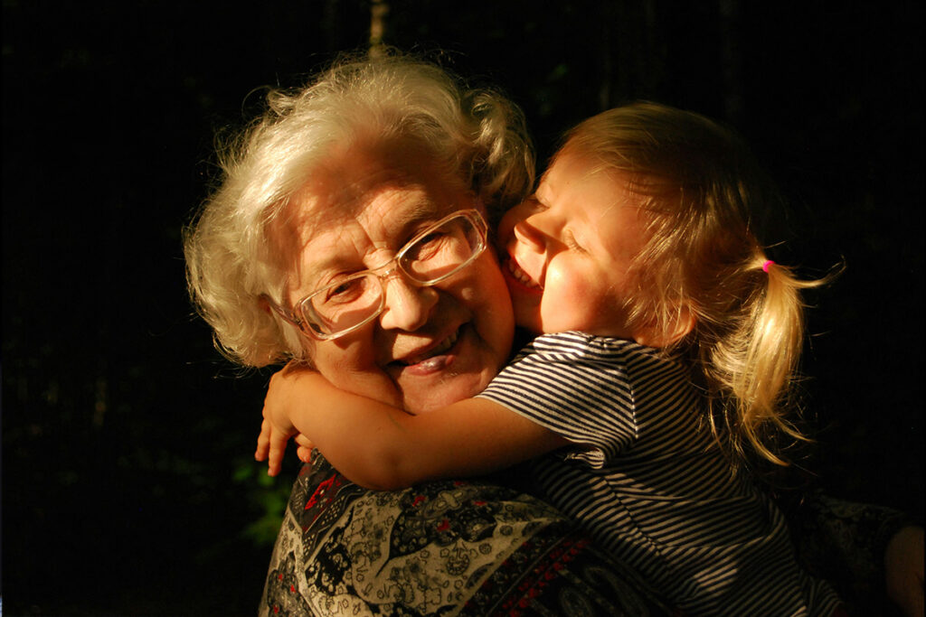 A small girl around 4 years old hugging her grandma and kissing her check. The Grandma has a sweet smile on her face.